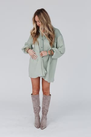 A woman with long, wavy blonde hair wears the Sunday Kind Of Love Dress in sage, pairing it with colorful bracelets and taupe knee-high suede boots. She stands against a white background, looking down and smiling.