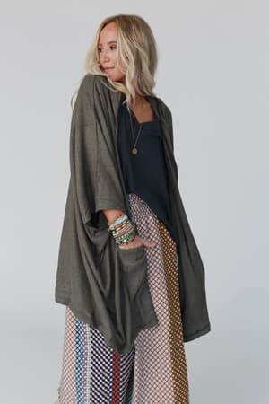 A woman with long blonde hair wears The Nest Boho Essential Hooded Ribbed Sweater in olive over a navy top and patterned wide-leg pants, accessorized with colorful bracelets and a gold pendant necklace against a light background.