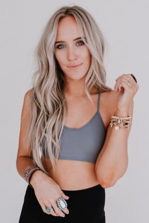 A woman with long, wavy blonde hair wears the Tattoo Bralette - Gray and black pants. She poses against a plain white background, showing off several bracelets and rings on her hand.