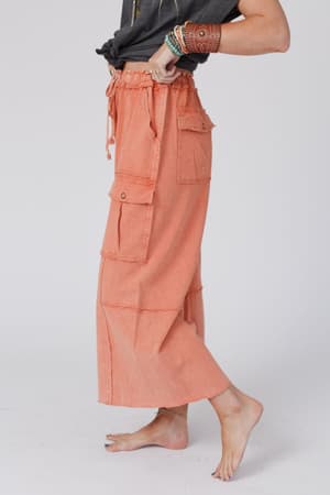 A person wears the Game On Cargo Wide Leg Pant in Burnt Orange with a black top, standing barefoot with one foot raised, showcasing stacked bracelets. The pants feature large pockets. The background is plain and light-colored.