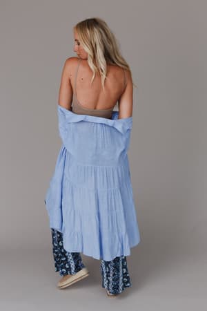 A woman with long blonde hair stands with her back to the camera, wearing the Dora Low Back Seamless Bralette in Mocha under a loose light blue tiered dress, patterned blue pants, and beige espadrilles, against a plain gray background.