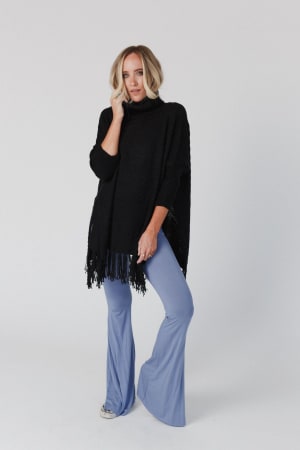 A blonde woman poses confidently against a white background, wearing the Paloma Poncho Sweater in black with an oversized silhouette and light blue flared pants, resting one hand on her face.