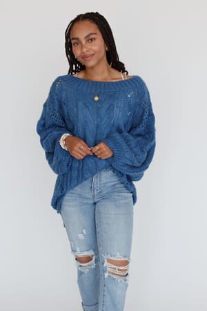 A woman with braided hair wears the Golden Hour Bubble Sleeve Sweater Dress in blue, paired with layered necklaces and bracelets. She stands against a plain white background, smiling slightly.