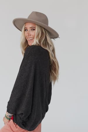 A woman with long blonde hair smiles over her shoulder, wearing The Nest Fly With Me Batwing Sleeve Knit Sweater in charcoal, a wide-brimmed beige hat, pink pants, and colorful bracelets. The background is a plain light color.