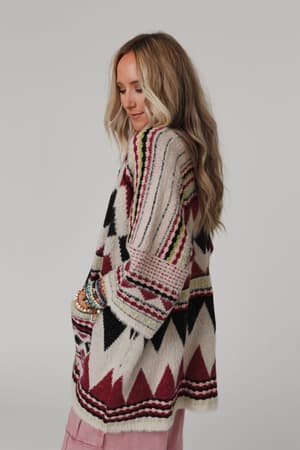 A woman with long, wavy blonde hair wears the Keep It Up Oversized Cardigan - Berry, featuring maroon, black, and cream geometric designs over pink pants. She stands sideways, looking down with hands in her pockets against a plain light background.
