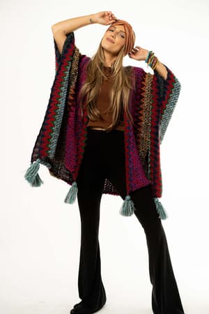 A woman models the vibrant Desert Dreams Crochet Ruana - Magenta with tassels, black flared pants, a brown top, rust headwrap, and layered bracelets against a white background—an ideal statement piece for any boho collection.