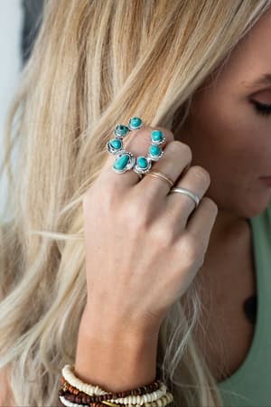A close-up of a blonde woman wearing the Celia Blossom Turquoise Ring in silver tone, a thin gold band, and beaded bracelets with natural-cut beads. She holds her hand near her face and wears a green top.