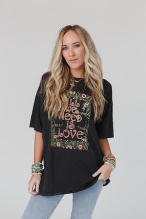 A woman with long, wavy blonde hair wears the Share The Love Graphic Tee in charcoal, paired with light blue jeans and colorful beaded bracelets, standing against a plain light background.