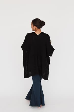 A woman with her hair in a bun stands facing away, wearing the So Deserving Knit Ruana in black and wide-leg dark blue jeans against a plain white background.