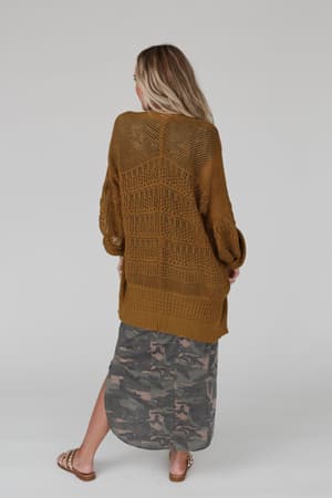 A woman with long blonde hair, facing away, wears the Brenden Knit Netted Cardigan in brown over a camouflage skirt and sandals, standing against a plain light background.