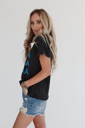 A woman with long, wavy blonde hair wears the Thunder Mama Vintage Graphic Tee in charcoal with distressed denim shorts, standing sideways and smiling over her shoulder against a plain white background.