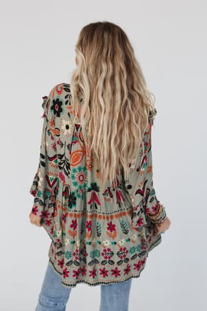 A woman with long, wavy blonde hair faces away wearing the Leona Floral Print Tunic in Sage, featuring a v-neckline and ruffled sleeves, paired with light blue jeans, set against a plain white background.