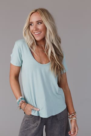 A smiling woman with long blonde hair wears the Scoop Neck Bralette Tee in Washed Seafoam, paired with gray pants and colorful beaded bracelets. She stands against a plain gray background with one hand in her pocket.