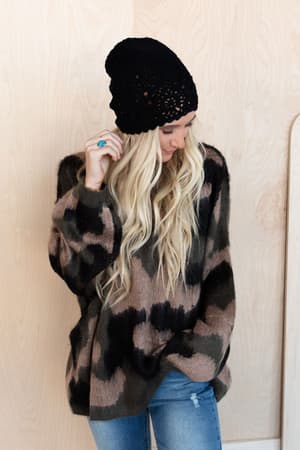 A woman with long blonde hair wears a black knit beanie, blue jeans, a turquoise ring, and the Wild Wanderer Sweater - Camo, standing against a light wooden background while looking down.