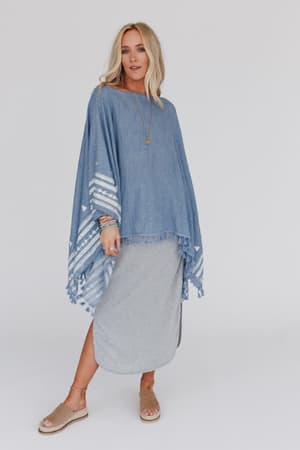 A woman with long blonde hair wears the Fallon Embroidered Sleeve Tassel Poncho in blue, styled over a gray skirt with layered bracelets, a necklace, and tan open-toe sandals. She stands against a plain white background.