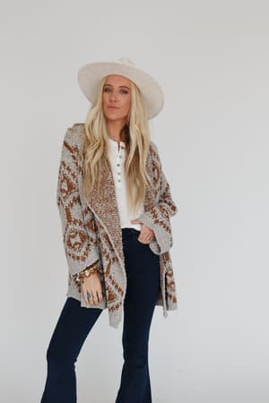 A woman with long blonde hair wears the Free to Roam Cardigan in gray over a white top, paired with a wide-brimmed white hat and dark flared jeans, standing against a plain white background.