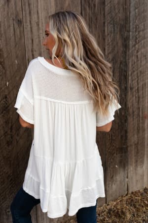 A woman with long, wavy blonde hair stands outdoors facing a wooden fence. She wears the More Than Meets The Eye Tunic Top in ivory with textured and mesh details, paired with dark jeans. Her face is turned to the side.