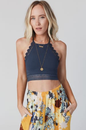 A woman with blonde wavy hair wears the Light Of The Moon Crochet Trim Bralette in blue gray and a yellow floral skirt with pockets, accessorized with layered necklaces, posing with her hands in her skirt pockets against a light background.