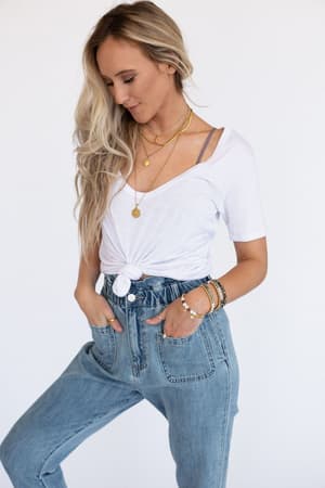 A woman with long blonde hair wears the Scoop Neck Bralette Tee in white, paired with high-waisted blue jeans and layered gold accessories. She looks down with her hands in her pockets against a plain white background.
