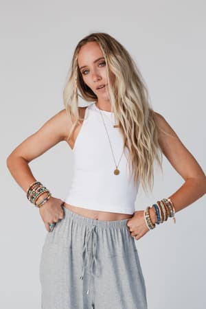 A woman with long, wavy blonde hair wears the High Neck Racerback Brami in white with gray drawstring pants, layered bracelets, and a necklace. She stands against a plain background with her hands on her hips.