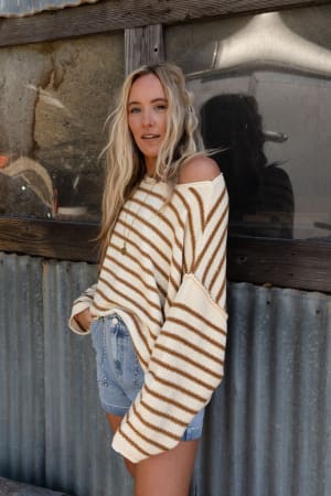 A woman with long blonde hair wears the All My Stripes Oversized Sweater in mustard with denim shorts, standing against a weathered metal and wood wall. The relaxed fit of her striped sweater adds to her casual vibe.