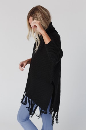 Wearing the Paloma Poncho Sweater - Black, a blonde model poses in light blue pants with her head down and hand raised against a plain light background.