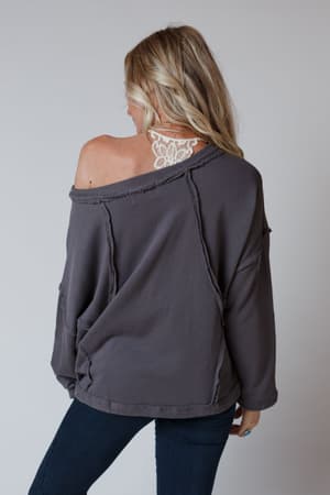 A woman with long blonde hair faces away, wearing the Bailey Oversized Raw Edge Top in charcoal over a white lace bralette and dark jeans, set against a plain light background.