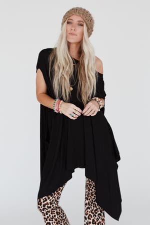 A woman with long blonde hair wears The Wren Tunic - Black, a chunky knit beanie, leopard print pants, and colorful bracelets while standing against a plain white background.