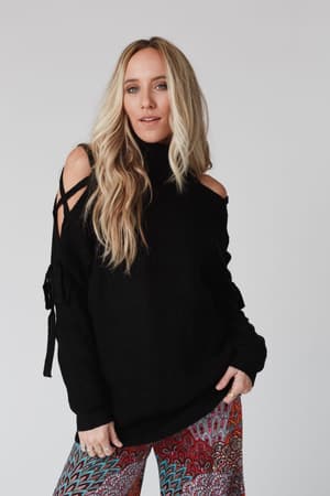 A woman with long blonde hair wears the Roxy Criss Cross Tie Shoulder Sweater in black, paired with colorful patterned pants, standing against a plain light background.