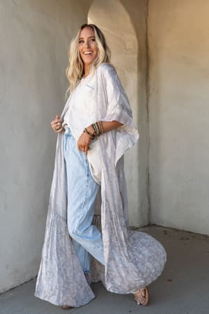 A woman with long blonde hair smiles while wearing the Paisley Tapestry Free Flow Duster Kimono in gray over a white top and light blue jeans, standing against a beige stucco archway with bracelets and sandals.