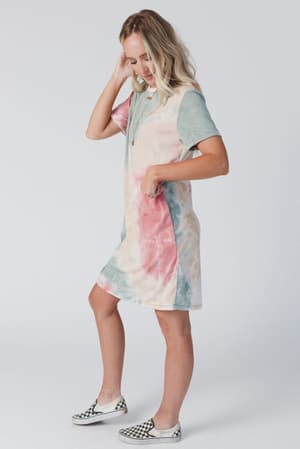 A woman wears The Nest On The Daily Oversized Slit Tee Dress - Tie Dye, featuring side slits. She stands sideways with one hand in her hair, the other in her pocket, styled with checkered slip-on sneakers and a necklace against a white background.