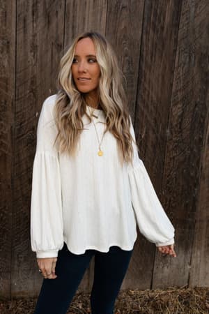 A woman with long, wavy blonde hair wears The Nest Sweet Heart Balloon Sleeve Knit Sweater in cream and dark jeans, standing before a wooden wall. She looks to the side with a gold necklace and slightly extends her right arm.