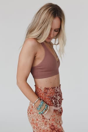 A woman with long blonde hair wears the Evermore Seamless Lace Racerback Bralette in mauve and high-waisted red floral pants, standing in profile with one hand in her pocket and several colorful beaded bracelets on her wrist.