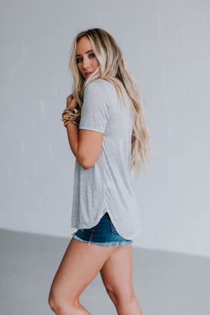 A woman with long blonde hair stands sideways, looking at the camera, wearing the Scoop Neck Bralette Tee - Gray and denim shorts against a plain light background.