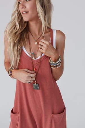A woman with long blonde hair wears a rust sleeveless dress over a white tank, accessorized with the Giselle Stone and Shell Boho Necklace - Turquoise and layered necklaces, her hands resting by her chest and pockets.