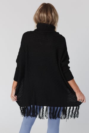 Seen from behind on a plain white background, a person with light brown hair wears the oversized Paloma Poncho Sweater in black with fringe along the hem, paired with light blue leggings and gently holding the sides.