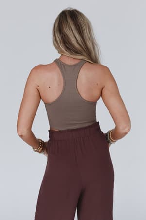 A woman with long blonde hair, seen from behind, wears the Lake Como High Neck Bralette in mocha and high-waisted dark brown pants. She has bracelets on both wrists and stands against a plain light gray background.