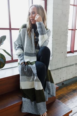 A woman with long, wavy blonde hair sits barefoot on wooden stairs by a large window, wearing black leggings and The Nest So Cozy Oversized Striped Knit Duster in Olive Multi.