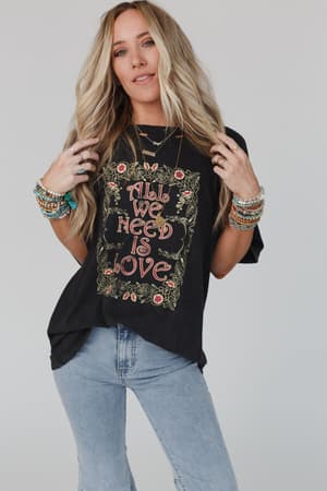 A woman with long, wavy blonde hair wears the Share The Love Graphic Tee in charcoal, styled with blue jeans and colorful beaded bracelets, standing against a plain light background.