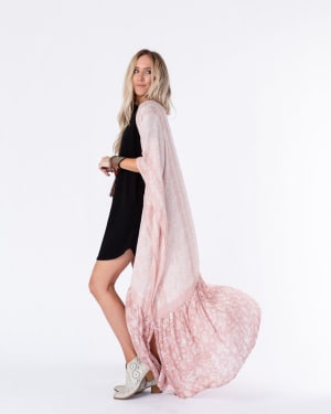A woman with long blonde hair wears a black dress, white ankle boots, and the Paisley Tapestry Free Flow Duster Kimono in Blush. She stands sideways against a plain white background, the kimono flowing gracefully behind her.