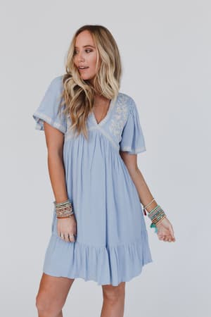 A woman with long, wavy blonde hair wears the Magdaline Mini Dress in blue, featuring short sleeves and a ruffle hem, accessorized with colorful bracelets, standing against a plain white background.