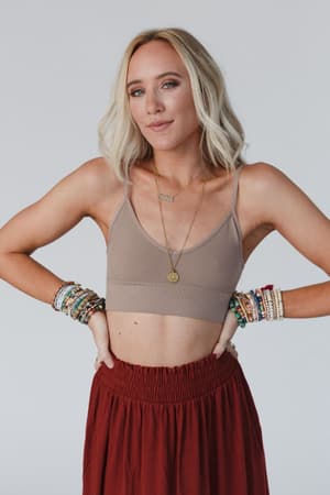 A woman with shoulder-length blonde hair wears the Dora Low Back Seamless Bralette in Mocha, a maroon skirt, layered necklaces, and colorful beaded bracelets, standing with her hands on her hips against a plain background.