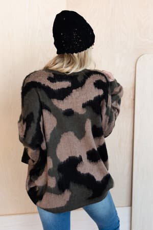 A person wearing a black knit beanie, light blue jeans, and the Wild Wanderer Sweater - Camo stands facing away from the camera against a light wood background.