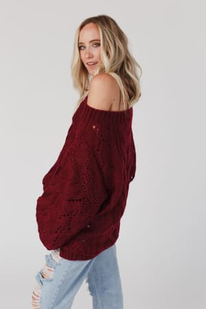 A woman with blonde hair wears the Golden Hour Bubble Sleeve Sweater Dress in burgundy, standing sideways and looking over her shoulder against a plain white background.