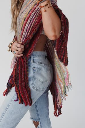 A person with long hair, wearing light blue ripped jeans and the colorful Madeleine Poncho - Multi, stands side-on to the camera, accessorized with bracelets and rings.