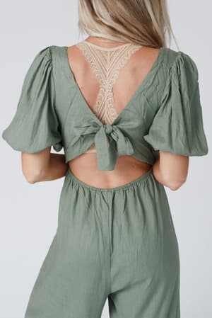 A woman wears the Weekender Puff Sleeve Jumpsuit in olive, featuring wide legs, a deep V-back, a waist bow tie, and beige lace detailing. The lightweight fabric offers comfort and style.