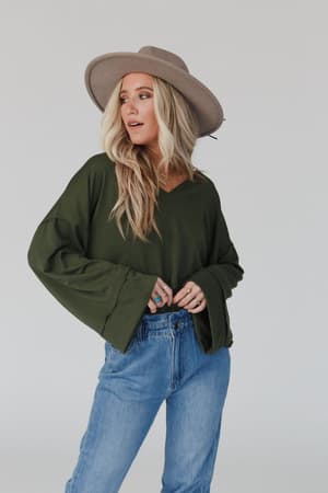 A woman in The Nest Love It V Neckline Sweater Top - Olive, a wide-brimmed tan hat, and blue jeans stands against a plain light gray background, looking to the side.