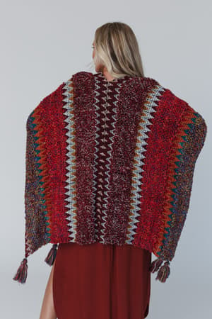 A woman with long blonde hair is seen from the back, wearing a knit Desert Dreams Crochet Ruana - Maroon over a burgundy dress, making it a perfect boho closet addition against a plain gray background.