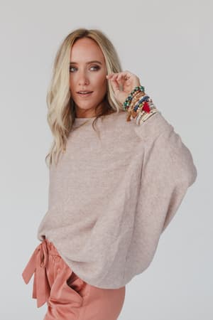A woman with long blonde hair models The Nest Fly With Me Batwing Sleeve Knit Sweater in Sand, paired with peach pants and colorful beaded bracelets, as she poses against a plain background looking slightly to the side.