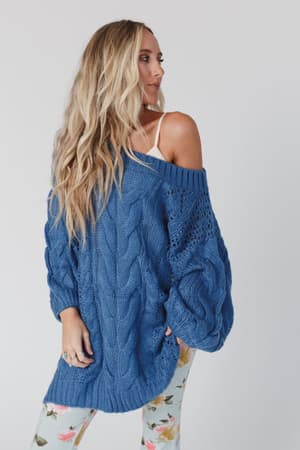 A woman with long blonde hair wears the Golden Hour Bubble Sleeve Sweater Dress in blue, styled over a white top and floral leggings. She looks to the side against a plain, light background.